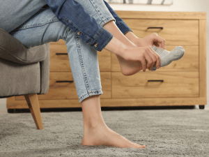 Person sits barefoot on a gray carpet, putting on a light gray sock near a wooden dresser.